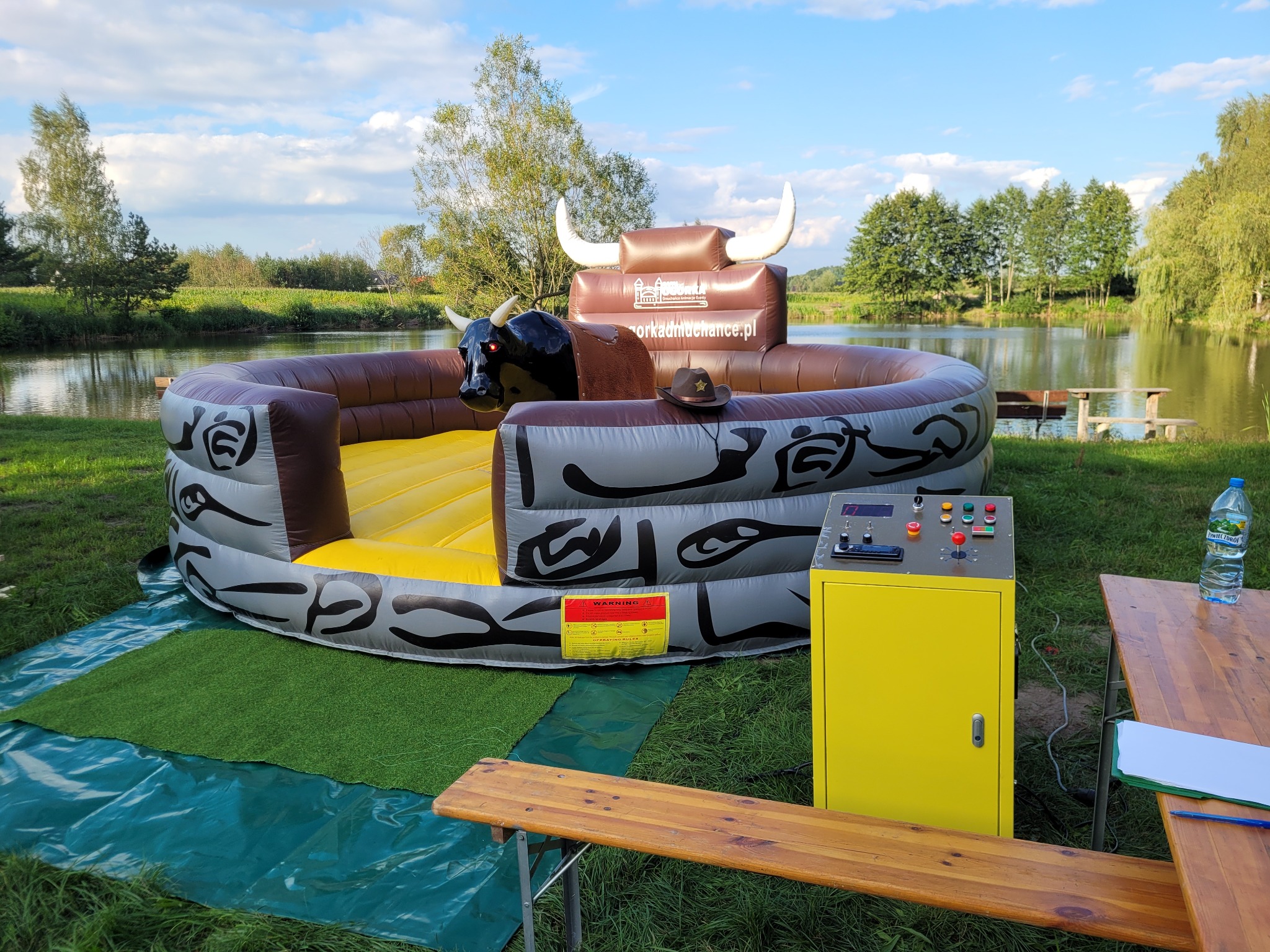 Nadmuchiwany byk rodeo z czarną głową, brązowym siodłem i kowbojskim kapeluszem, ustawiony na trawie obok jeziora, z żółtym panelem sterowania i butelką wody na stole w tle.