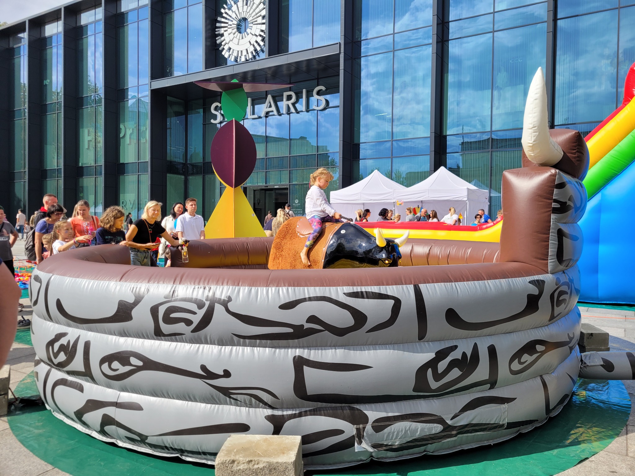 Dziewczynka w białej bluzce jeździ na dmuchanym byku na tle budynku Solaris, widoczny dmuchany zamek i grupa ludzi w tle.