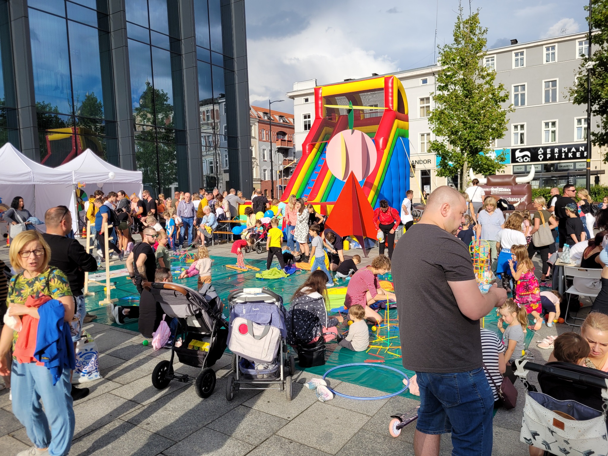 Tłum ludzi na placu miejskim podczas wydarzenia plenerowego, z dmuchanym zamkiem w tle i dziećmi bawiącymi się na macie edukacyjnej.