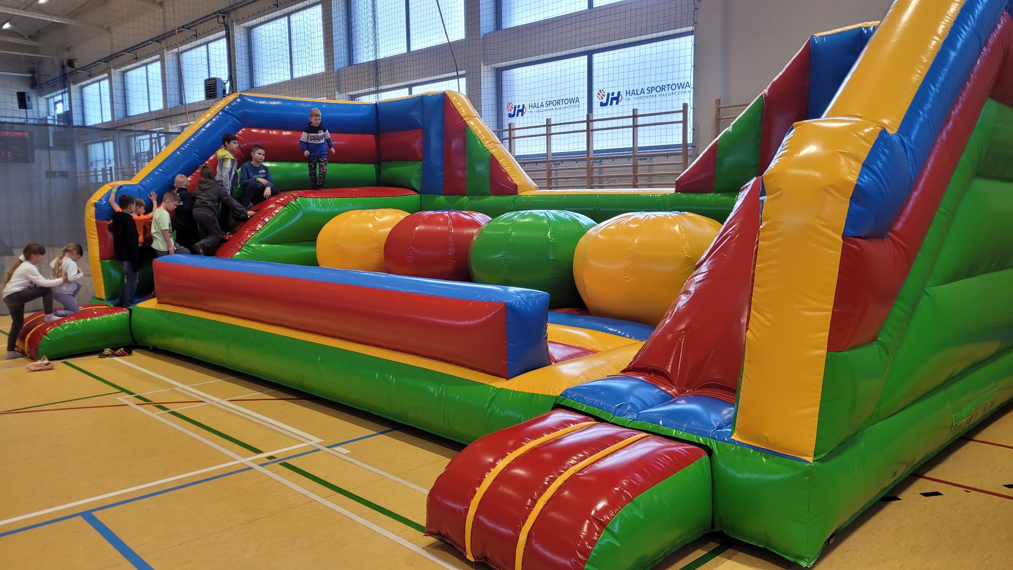 Duży, kolorowy dmuchany tor przeszkód z dziećmi w hali sportowej z parkietem i oknami w tle.
