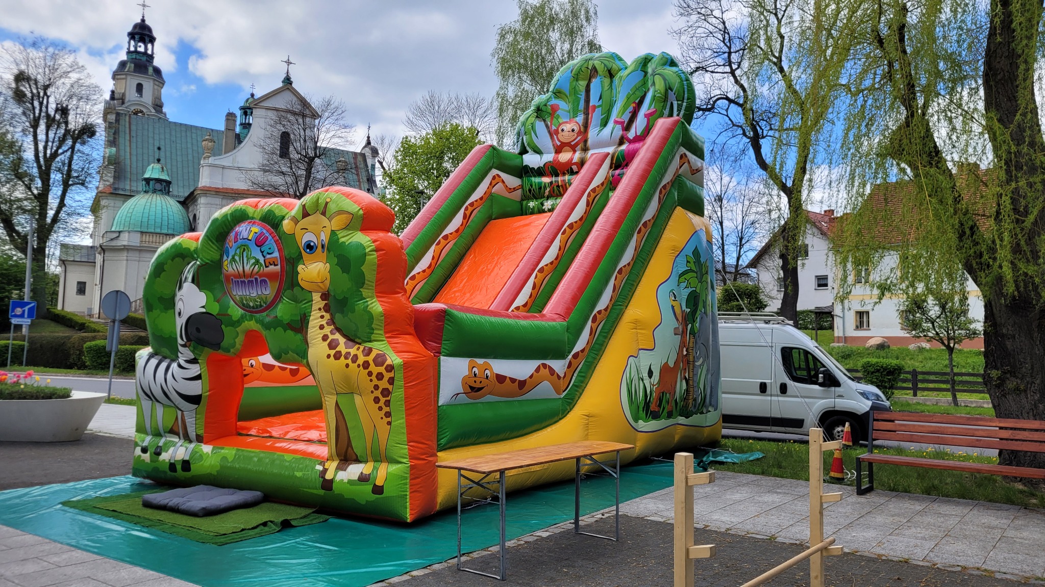 Kolorowa dmuchana zjeżdżalnia z motywem dżungli, zebrą, żyrafą i małpką, ustawiona na chodniku przed kościołem i zaparkowanym białym vanem.