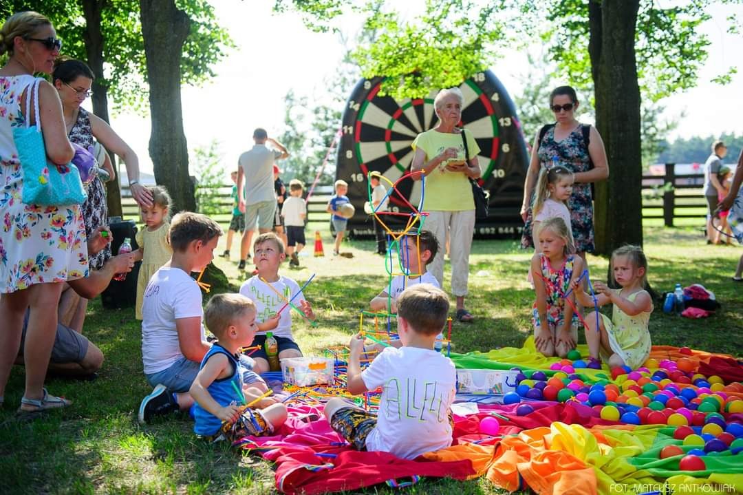 Grupa dzieci siedzi na kocu bawiąc się kolorowymi patyczkami i kulkami, w tle dmuchana tarcza do darta i dorośli