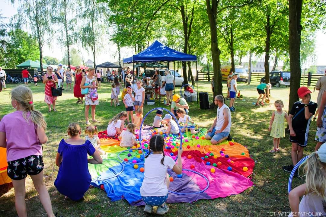 Grupa dzieci bawi się na kolorowej płachcie rozłożonej na trawie, otoczonej kolorowymi piłkami i obręczami hula-hop, w tle widoczni dorośli i namioty.