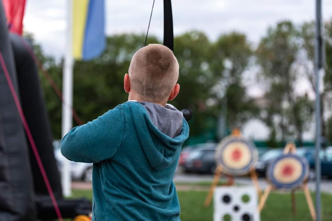 Chłopiec z krótkimi włosami, ubrany w turkusową bluzę, naciąga łuk na tle dmuchanego zamku i tarcz strzelniczych podczas imprezy plenerowej.