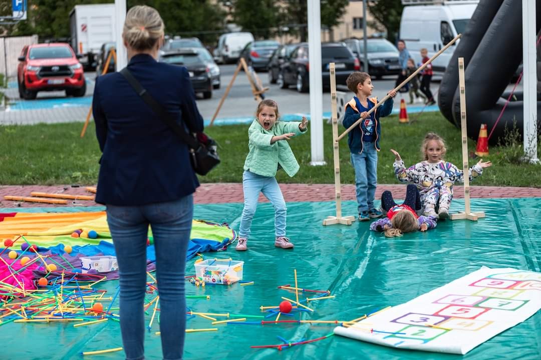 Dzieci bawią się na zielonej płachcie z grą w klasy i konstrukcjami z patyków, pod nadzorem kobiety, w tle dmuchana atrakcja.