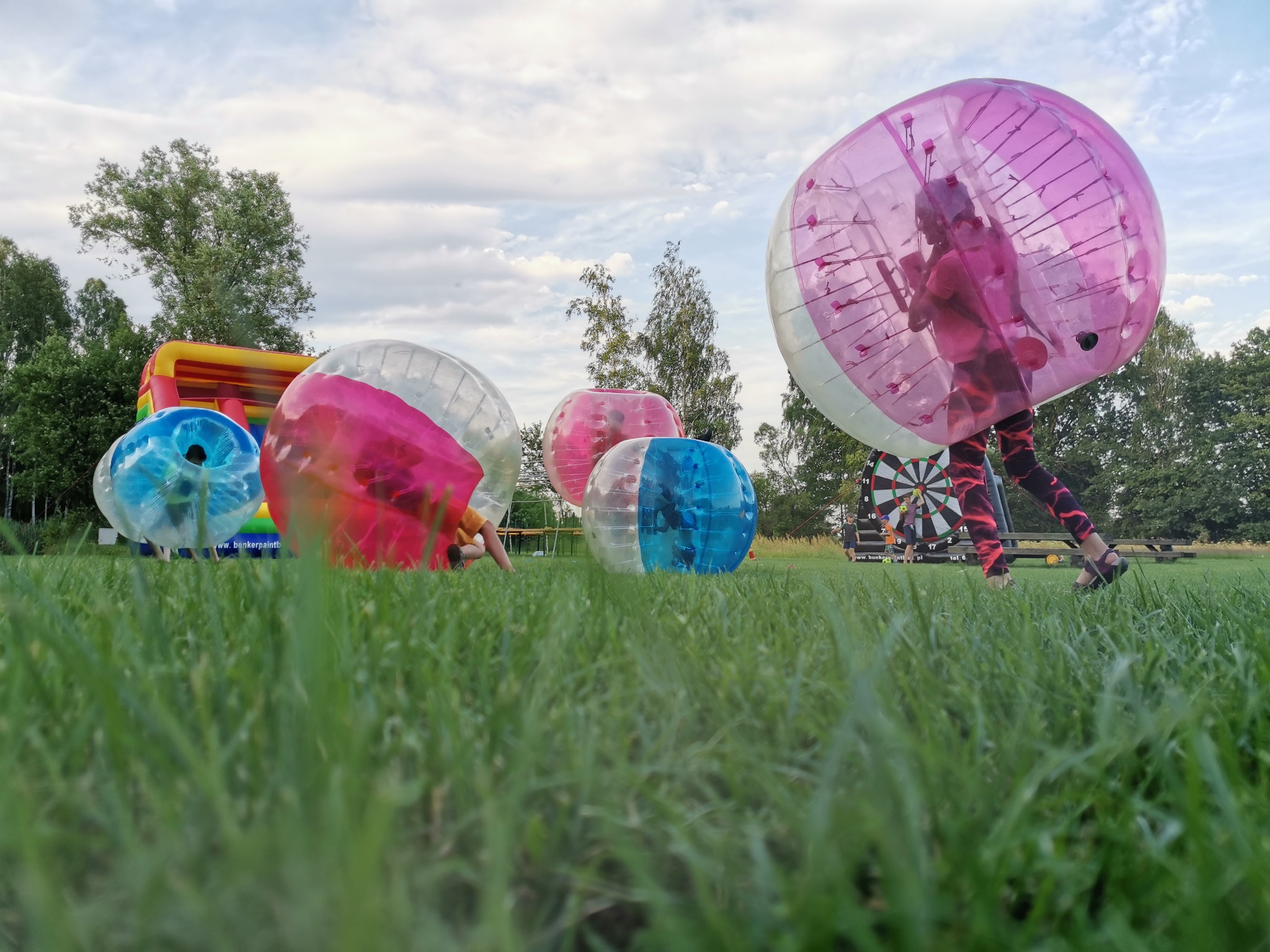 Osoba wewnątrz dużej, różowej kuli dmuchanej na trawiastym terenie, w tle inne kule, dmuchany zamek i tarcza do darta.