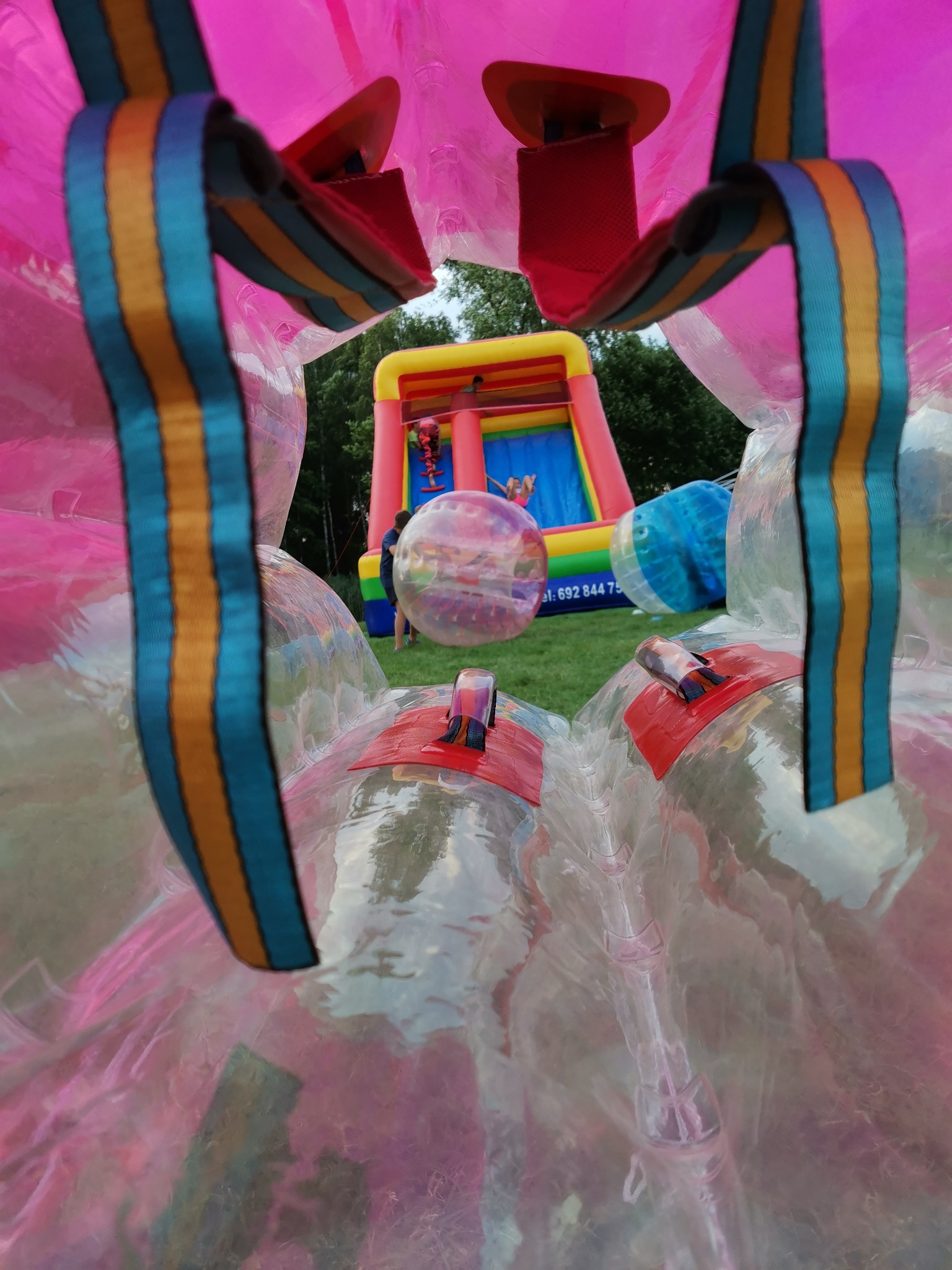 Widok z wnętrza dmuchanej kuli zorbingowej na kolorowy dmuchany zamek ze zjeżdżalnią i bawiące się dzieci na trawie.