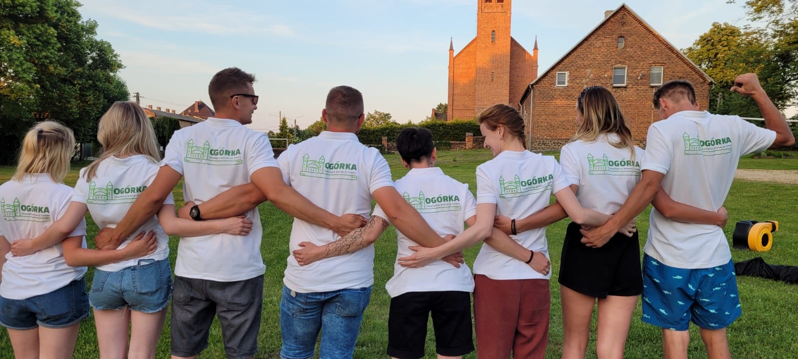 Grupa osób w białych koszulkach z logo firmy eventowej 'Ogórka' stojąca tyłem do kamery, obejmując się ramionami na tle ceglanego budynku i kościoła w promieniach zachodzącego słońca.