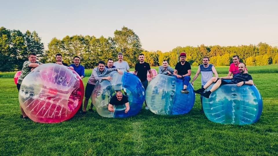 Grupa mężczyzn pozuje na trawie wewnątrz i obok kolorowych, przezroczystych kul zorbingowych na tle drzew i nieba.