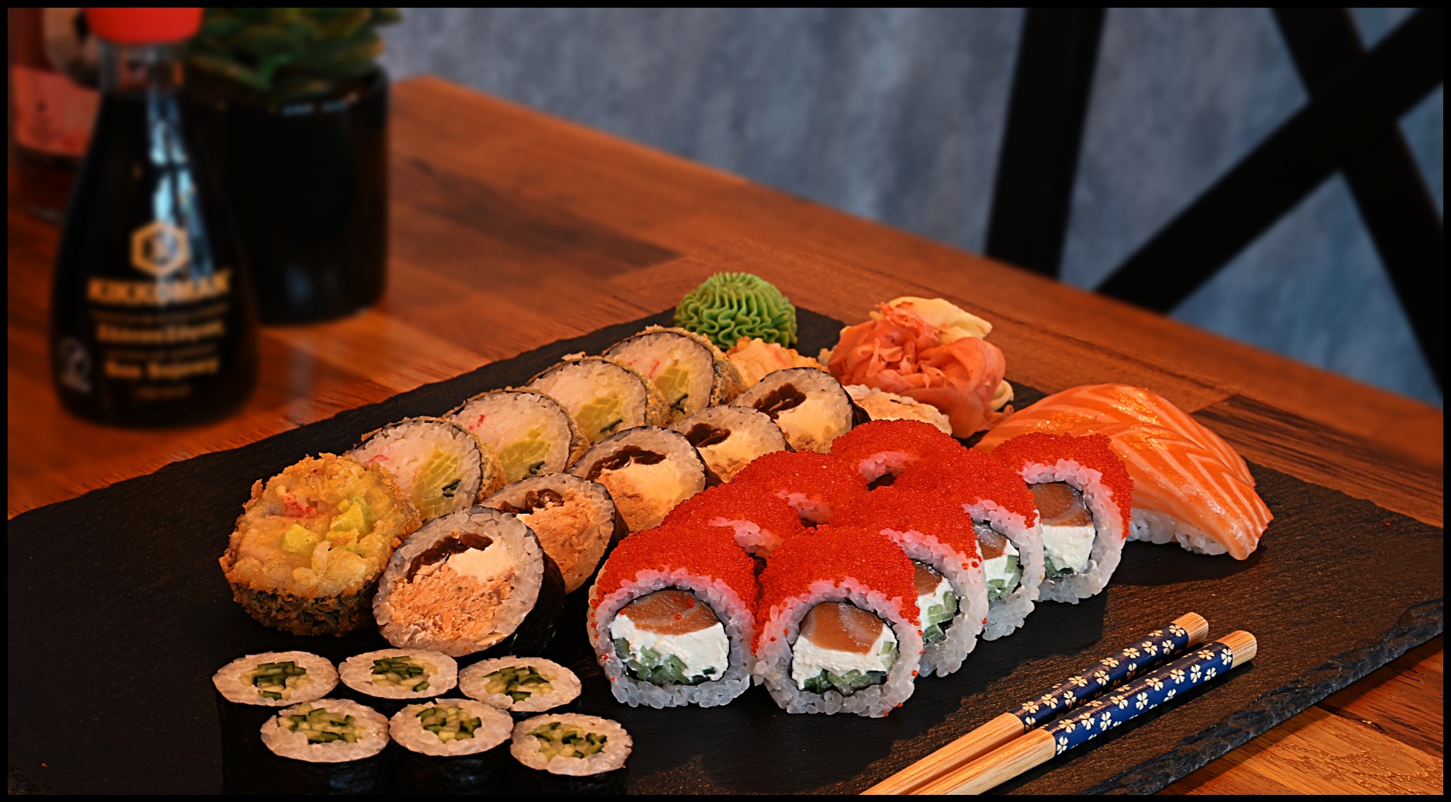 Zestaw różnorodnych rolek sushi na czarnym kamiennym talerzu, z pałeczkami w niebieskie kwiaty, sosem sojowym Kikkoman i rośliną w tle.