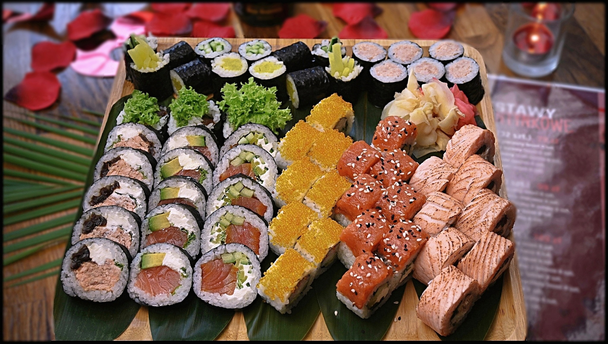 Zestaw różnorodnych rolek sushi ułożonych na drewnianej desce z liśćmi, w tle płatki róż i świeca, widoczny również fragment menu.