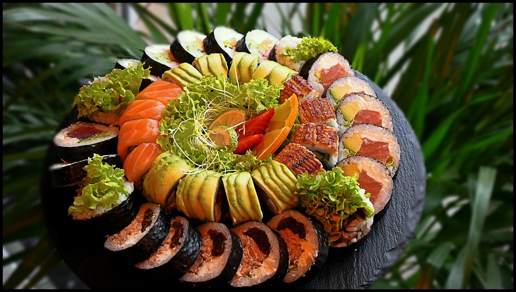 Dekoracyjny talerz z różnorodnymi rolkami sushi: łosoś, węgorz, awokado, tuńczyk, udekorowany świeżymi warzywami i cytrusami na tle roślin.