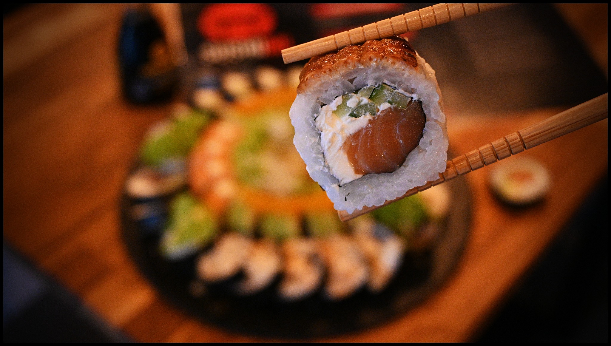 Pojedyncza rolka sushi z łososiem, ogórkiem i kremowym serem, trzymana pałeczkami na tle talerza z różnymi rodzajami sushi.