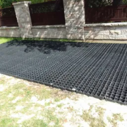 Czarna, plastikowa kratka trawnikowa ułożona na piasku, widoczny fragment kamiennego ogrodzenia w tle.