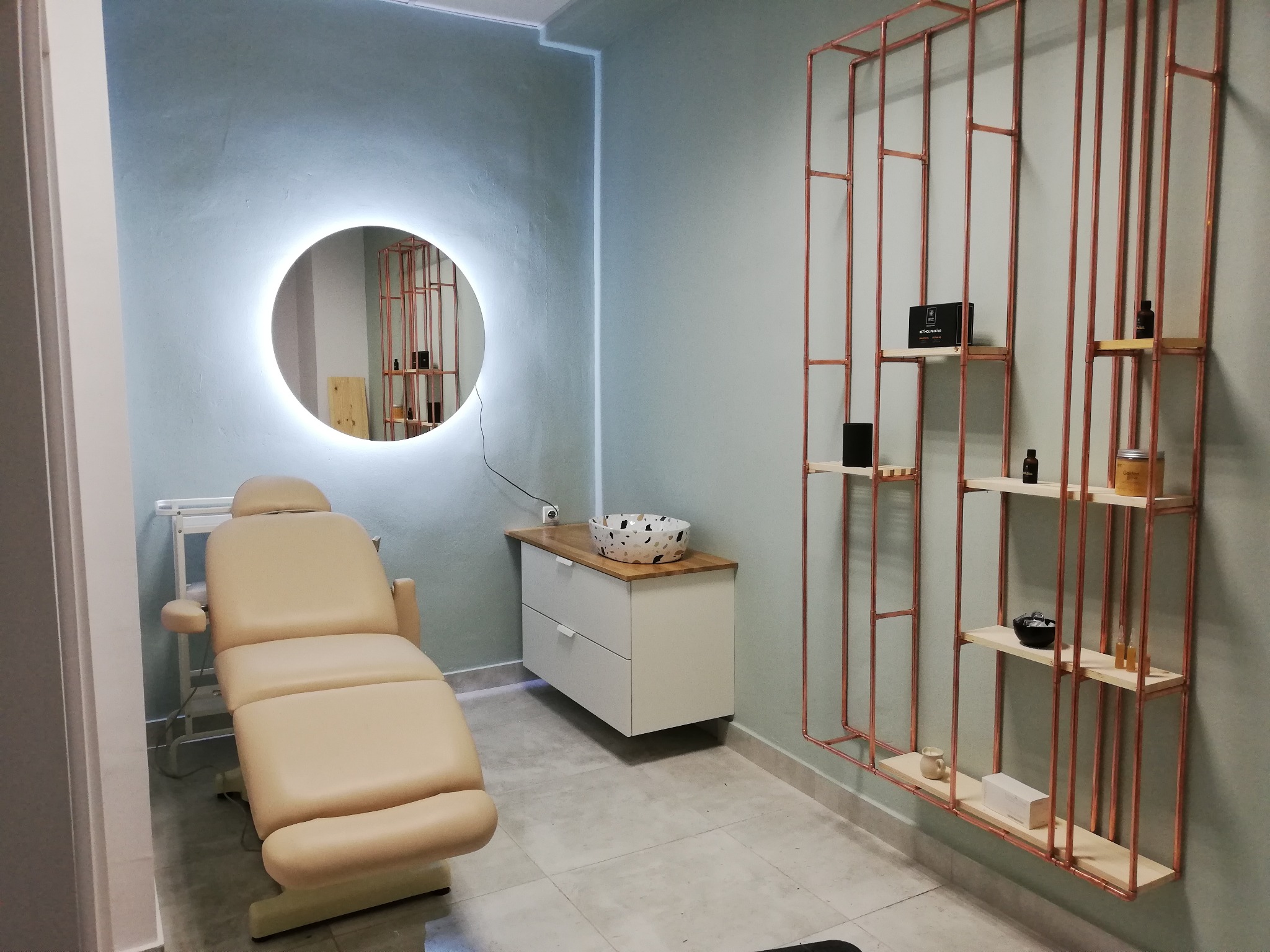 Jasny gabinet kosmetyczny z beżowym fotelem do zabiegów, okrągłym lustrem z podświetleniem i miedzianym regałem na kosmetyki