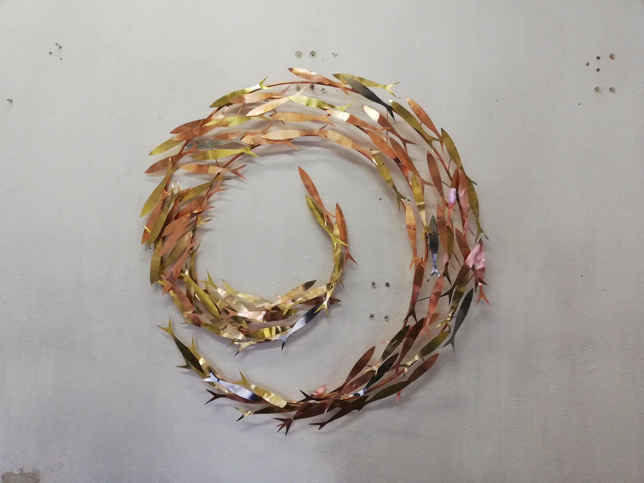 Metalowa dekoracja ścienna w kształcie spirali z ryb w odcieniach złota, miedzi i srebra, zamontowana na szarej ścianie.