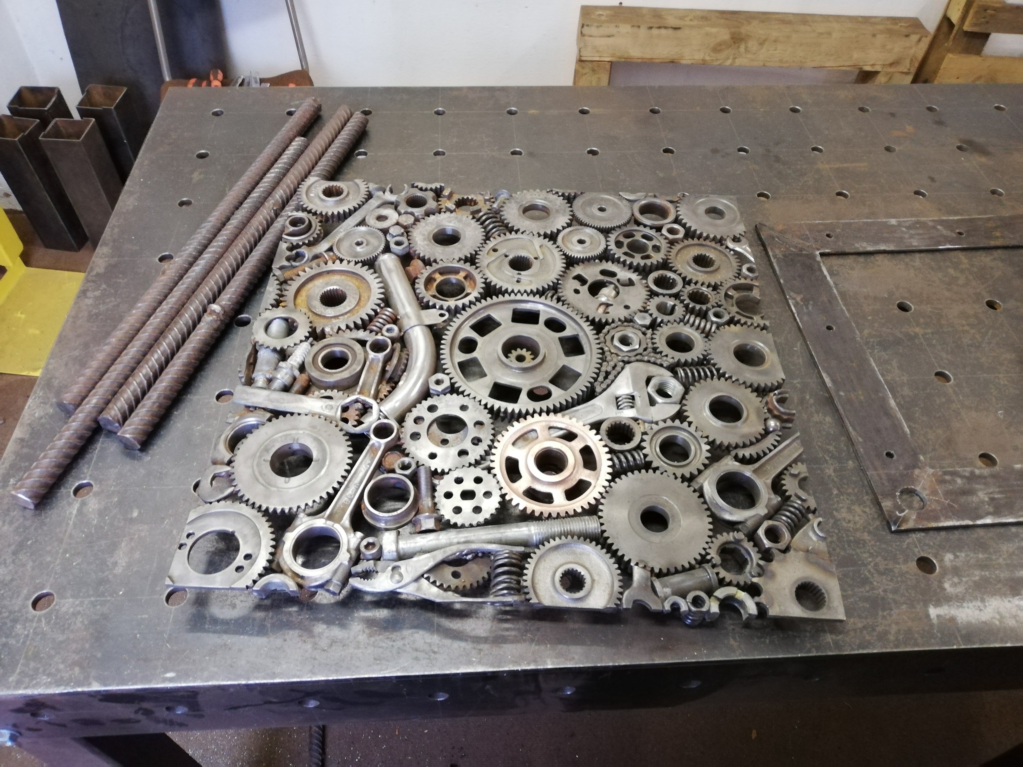 Metalowa kompozycja artystyczna z zębatek, sprężyn i innych elementów metalowych na perforowanym stole warsztatowym, obok leżą pręty zbrojeniowe i metalowe profile.