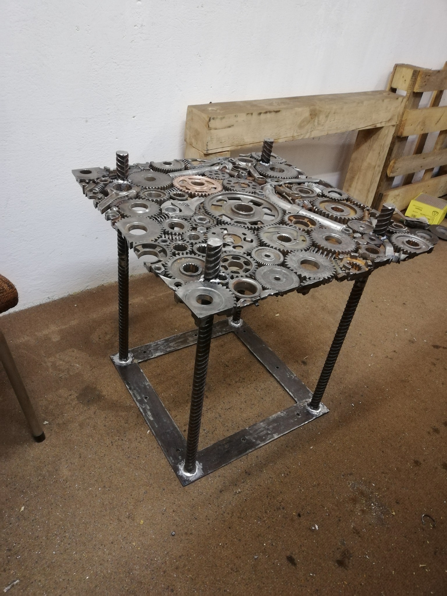 Stolik kawowy wykonany ze spawanych metalowych elementów, w tym kół zębatych i prętów żebrowanych, w surowym, industrialnym stylu.