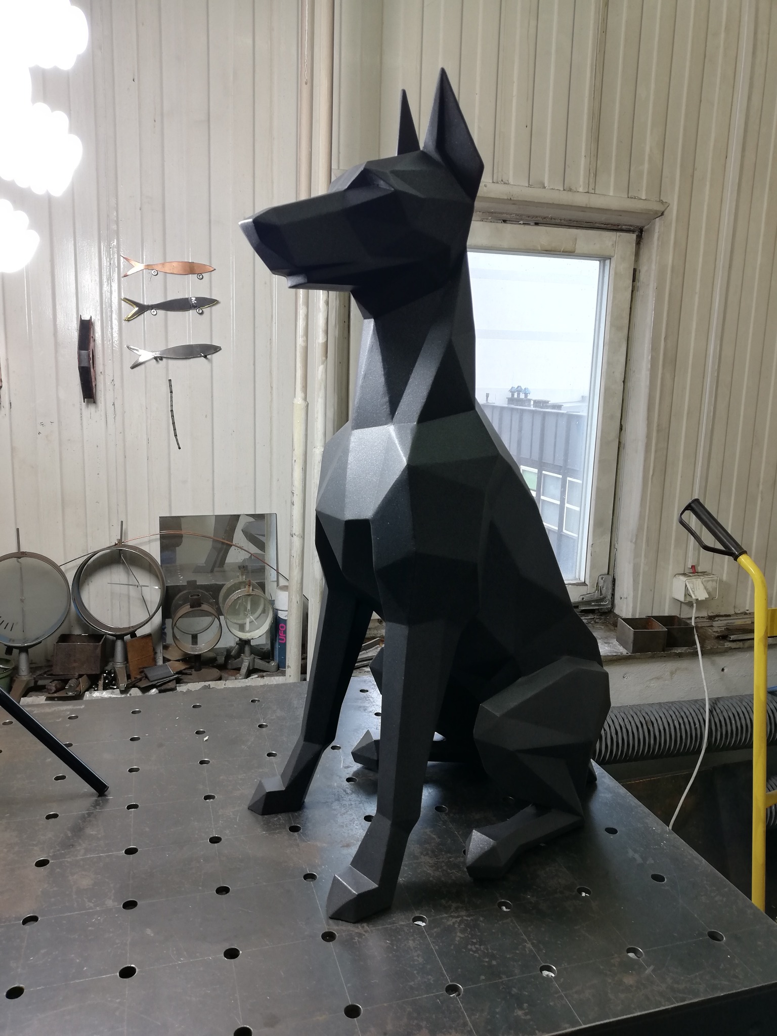 Geometryczna, czarna rzeźba psa na metalowym stole warsztatowym; w tle okno i metalowe ściany z wiszącymi dekoracjami ryb.