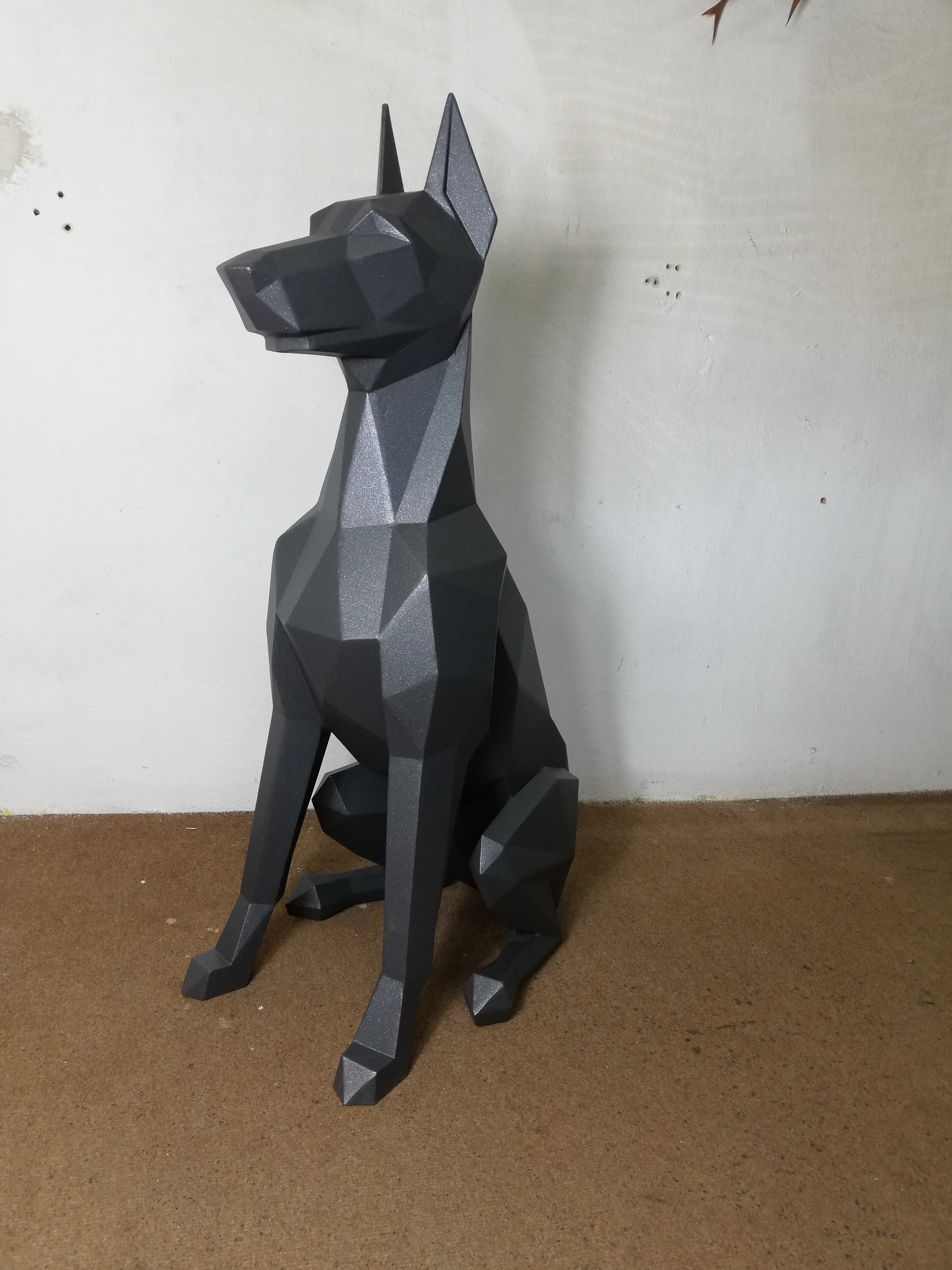 Geometryczna, metalowa rzeźba psa w kolorze grafitowym, siedząca na brązowym podłożu, na tle białej ściany z widocznymi śladami po wierceniu.