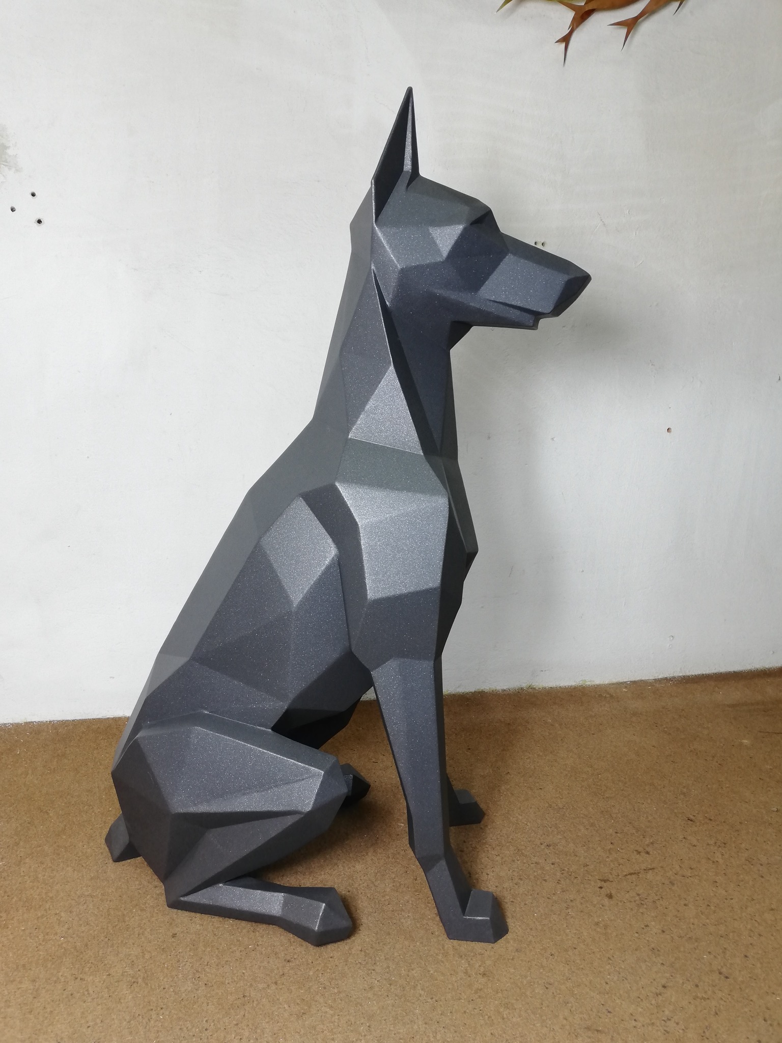 Geometryczna rzeźba psa w kolorze grafitowym, wykonana z metalu, siedząca na brązowej powierzchni, z widoczną białą ścianą w tle.