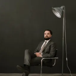 Portret brodatego mężczyzny w szarym tweedowym garniturze i krawacie w kratę, siedzącego na chromowanym krześle obok szarej lampy podłogowej na tle ciemnoszarej ściany.