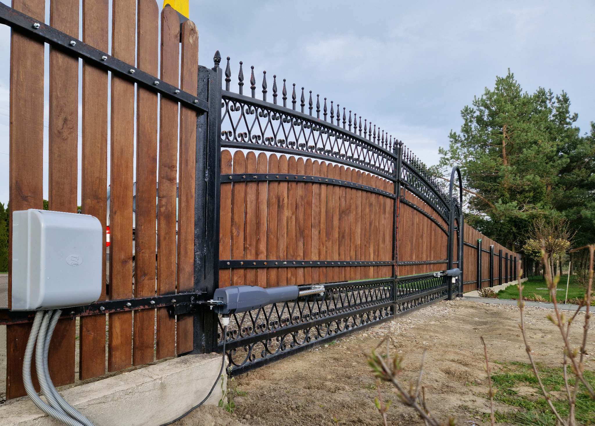 Dwuskrzydłowa brama wjazdowa z drewnianym wypełnieniem i ozdobnymi elementami kutymi, wyposażona w automat do bram z szarą obudową sterującą.