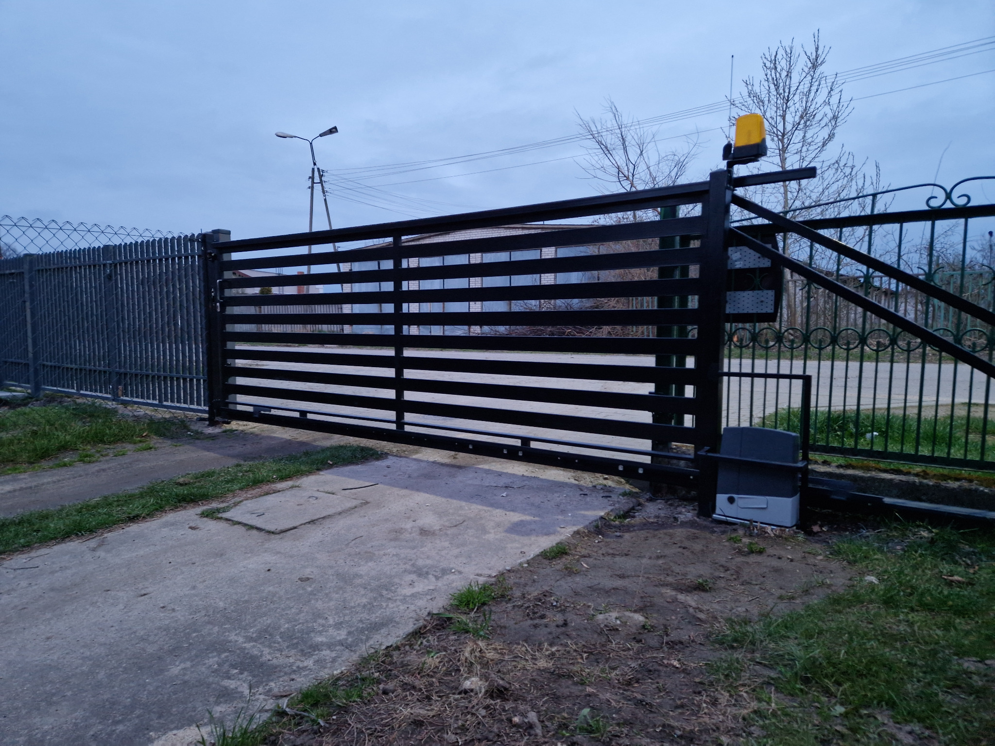 Przesuwna brama wjazdowa z automatyką, czarna, poziome profile, widoczny napęd i lampa ostrzegawcza, na tle niebieskiego nieba.