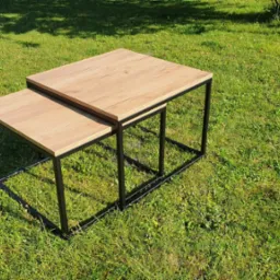 Dwa stoliki kawowe z drewnianymi blatami i czarnymi metalowymi nogami, ustawione jeden na drugim na trawniku w słoneczny dzień.