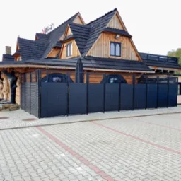 Ciemna, metalowa przesłona na tle drewnianego budynku w stylu góralskim z rzeźbami przed wejściem, widok z parkingu z kostki brukowej.