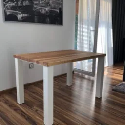 Stół na białych, kwadratowych nogach z blatem z jasnego drewna, stojący na brązowym parkiecie w pokoju z obrazem miasta w czarno-białej ramie.