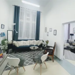 Jasne, przestronne wnętrze poczekalni z turkusową sofą z poduszkami futerkowymi, stolikiem kawowym, krzesłami z futerkowymi siedziskami i dywanem w geometryczny wzór, udekorowane roślinami...