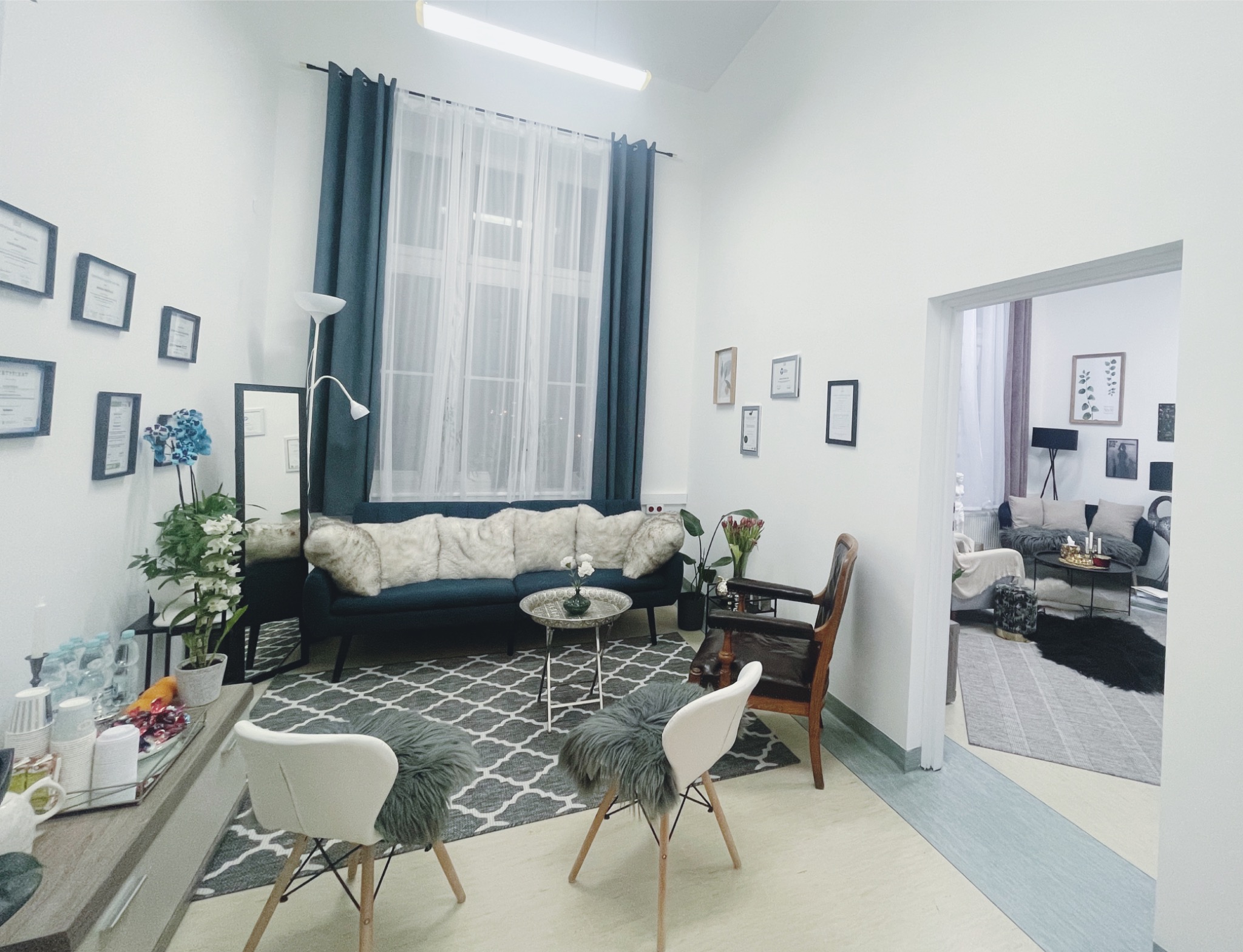 Jasne, przestronne wnętrze poczekalni z turkusową sofą z poduszkami futerkowymi, stolikiem kawowym, krzesłami z futerkowymi siedziskami i dywanem w geometryczny wzór, udekorowane roślinami...