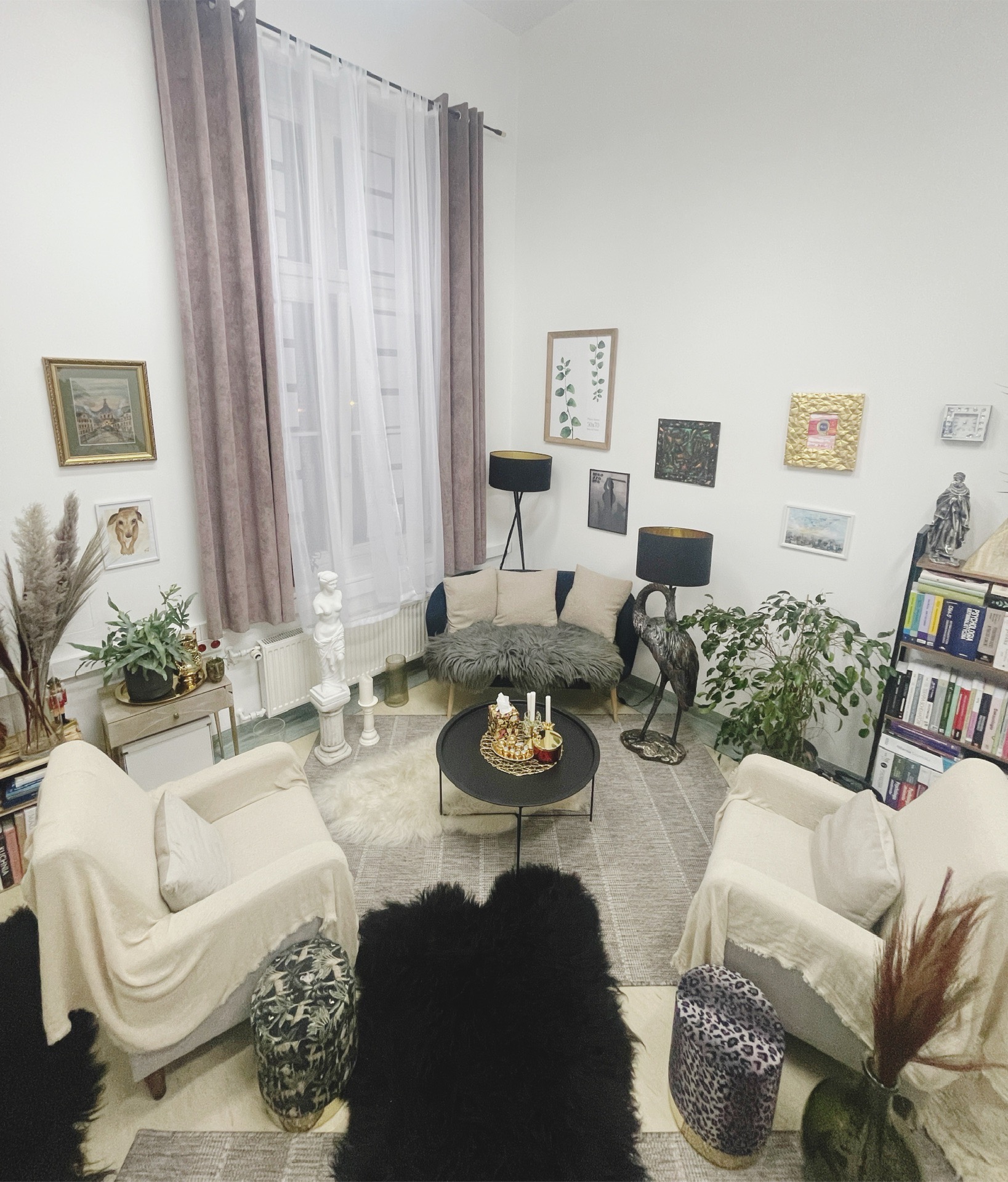 Jasny pokój z dwoma fotelami, sofą, stolikiem kawowym z dekoracjami, dywanami, lampami i półką z książkami tworzący przytulną przestrzeń do rozmów.