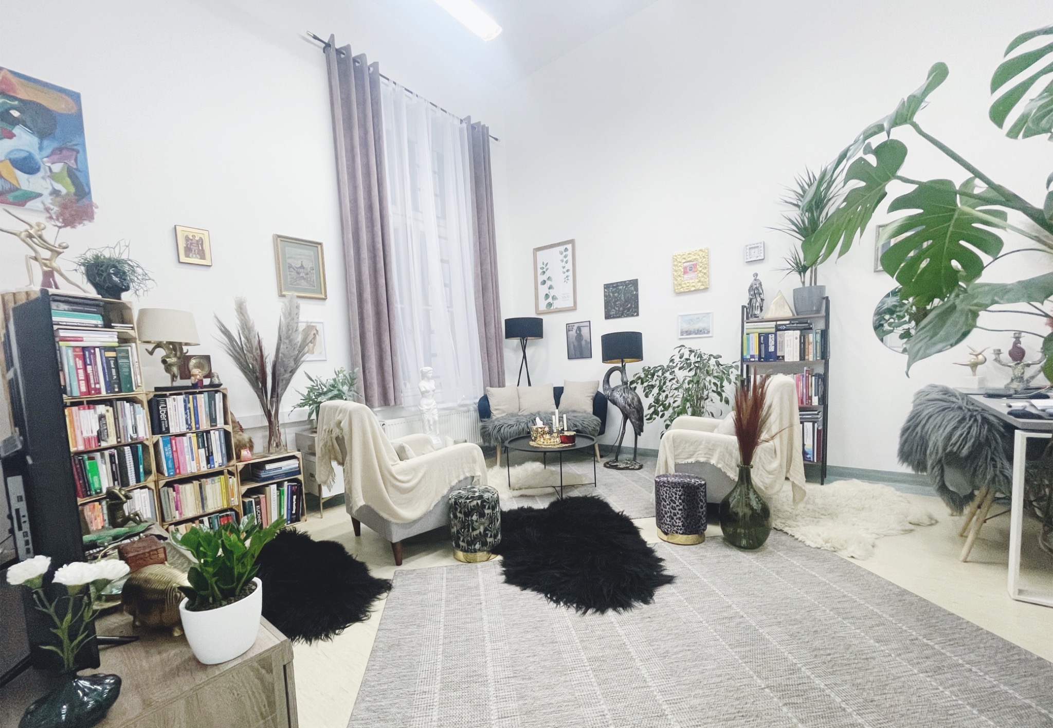 Jasne, przestronne wnętrze gabinetu z elementami wystroju: wygodne fotele z narzutami, stolik kawowy z dekoracjami, regały z książkami, rośliny doniczkowe i dywany w różnych stylach.