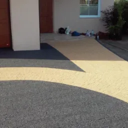 Ułożona kostka brukowa w dwóch odcieniach szarości i beżu tworząca podjazd do garażu i wejścia do domu. Na podjeździe widoczne ślady po pracy. W tle budynek z drewnianą bramą garażową i oknem.