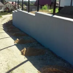 Wzniesiony mur oporowy z wystającymi słupkami, przygotowany do montażu ogrodzenia panelowego, widoczne pryzmy piasku wzdłuż ścieżki, domy w tle.