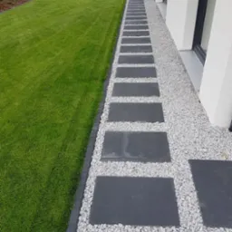 Ścieżka z prostokątnych płyt w ogrodzie, obłożona drobnym białym grysem, obrzeże z ciemnej kostki, trawnik po lewej, elewacja budynku po prawej.