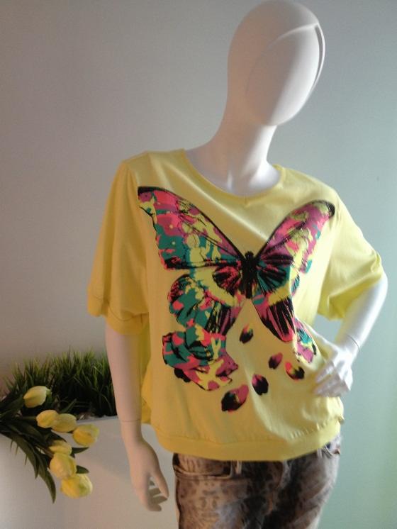 Żółta bluzka z krótkim rękawem prezentowana na manekinie, ozdobiona dużym, kolorowym motylem w odcieniach różu, zieleni i czerni, w tle bukiet żółtych tulipanów.