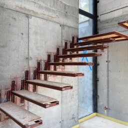 WeldOne - Montaż ażurowych schodów metalowych z drewnianymi stopniami na surowej betonowej ścianie, widoczne mocowania i przewody instalacyjne.