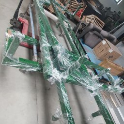 WeldOne - Elementy konstrukcji stalowej w kolorze zielonym, owinięte folią, przygotowane do transportu w Zielonce, widoczne w tle palety i wózek widłowy.