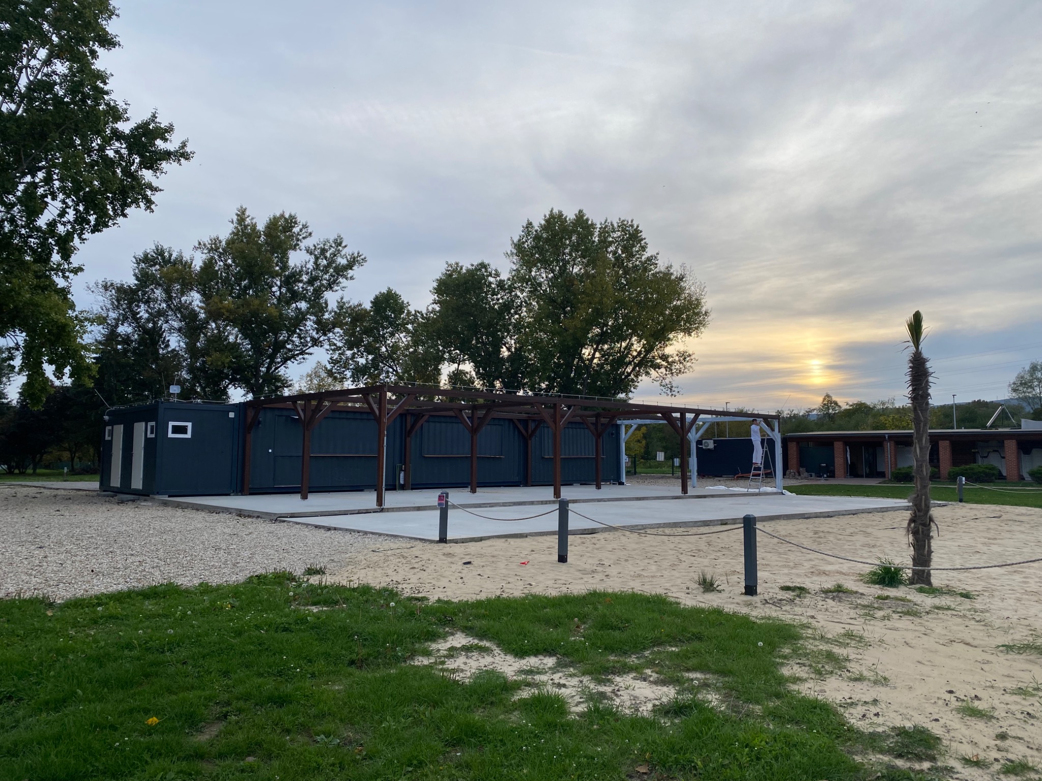 Konstrukcja z kontenerów i drewnianej pergoli na betonowej wylewce, otoczona piaskiem i trawą, w trakcie budowy na zewnątrz.