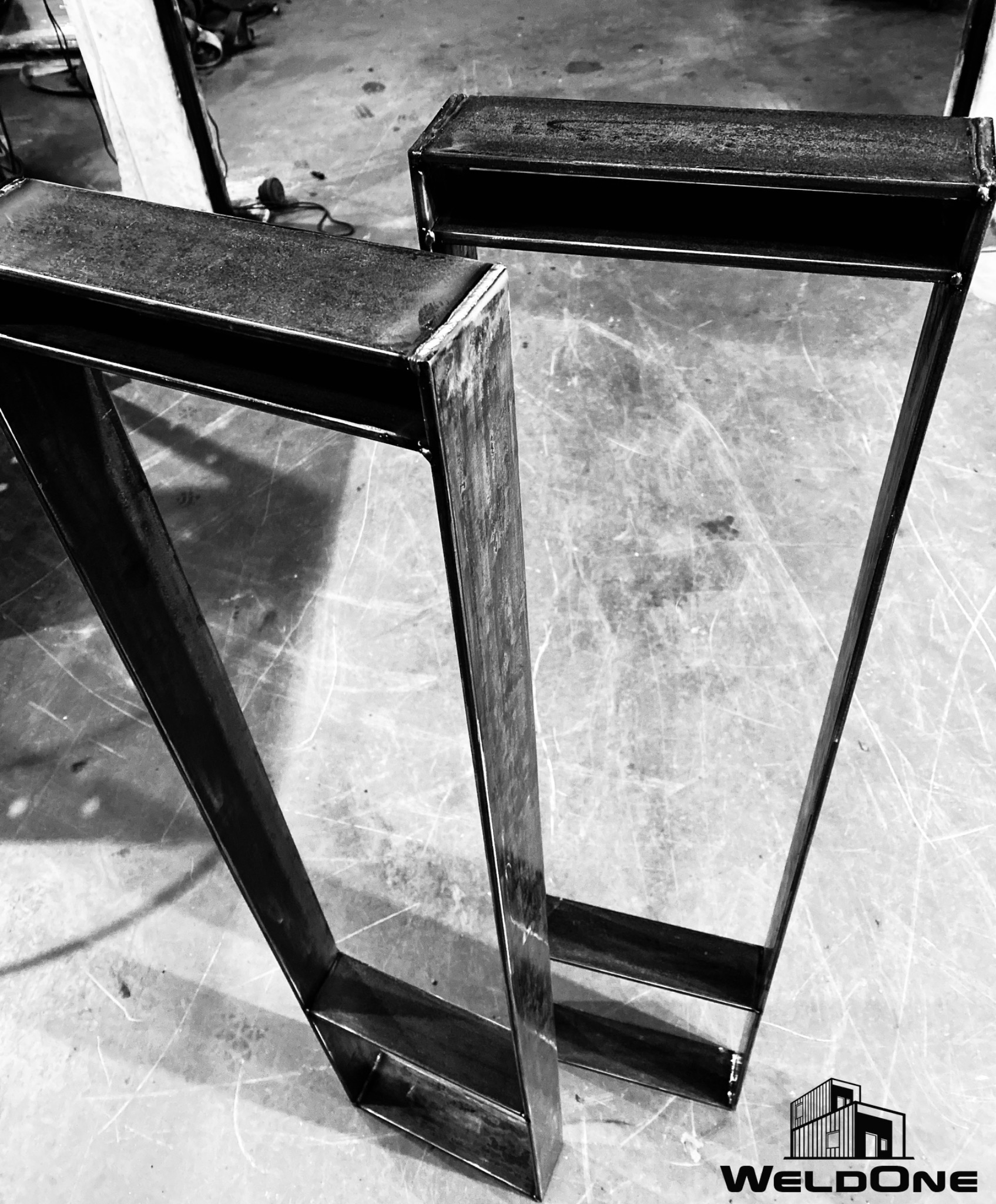 Dwie surowe, spawane ramy z metalowych profili o prostokątnym przekroju, leżące na posadzce warsztatu, widoczne ślady spawania i obróbki metalu, w tle narzędzia.