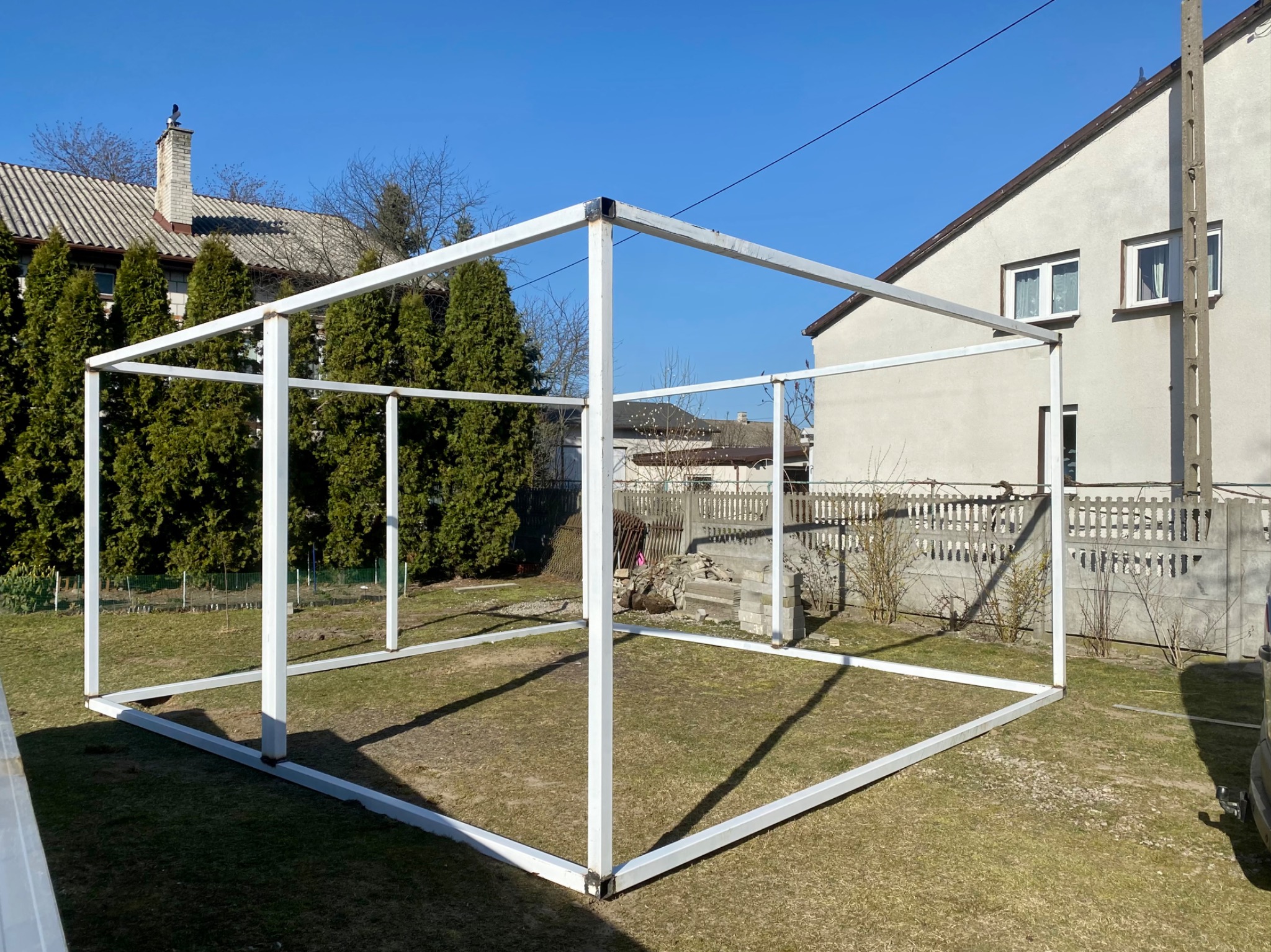 Szkielet metalowej konstrukcji w ogrodzie, spawane profile pomalowane na biało, przygotowane pod budowę hali lub altany, widoczne spawy na łączeniach.