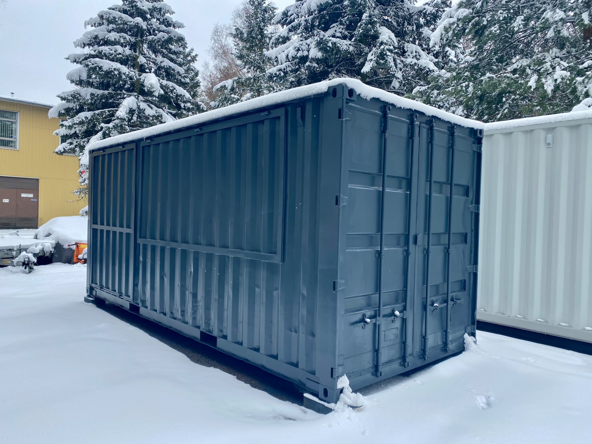Szary kontener morski pokryty śniegiem, stojący na zasypanym śniegiem terenie, z widocznymi drzewami iglastymi również pod śniegiem w tle.