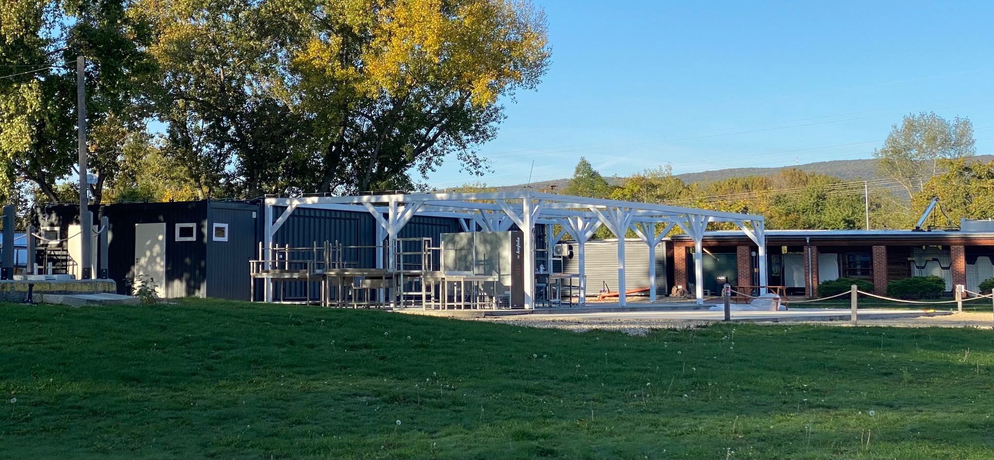 Konstrukcja z metalowych kontenerów i drewnianej pergoli w trakcie budowy, widoczne elementy stalowe i drewniane, trawnik z przodu, drzewa i góry w tle.