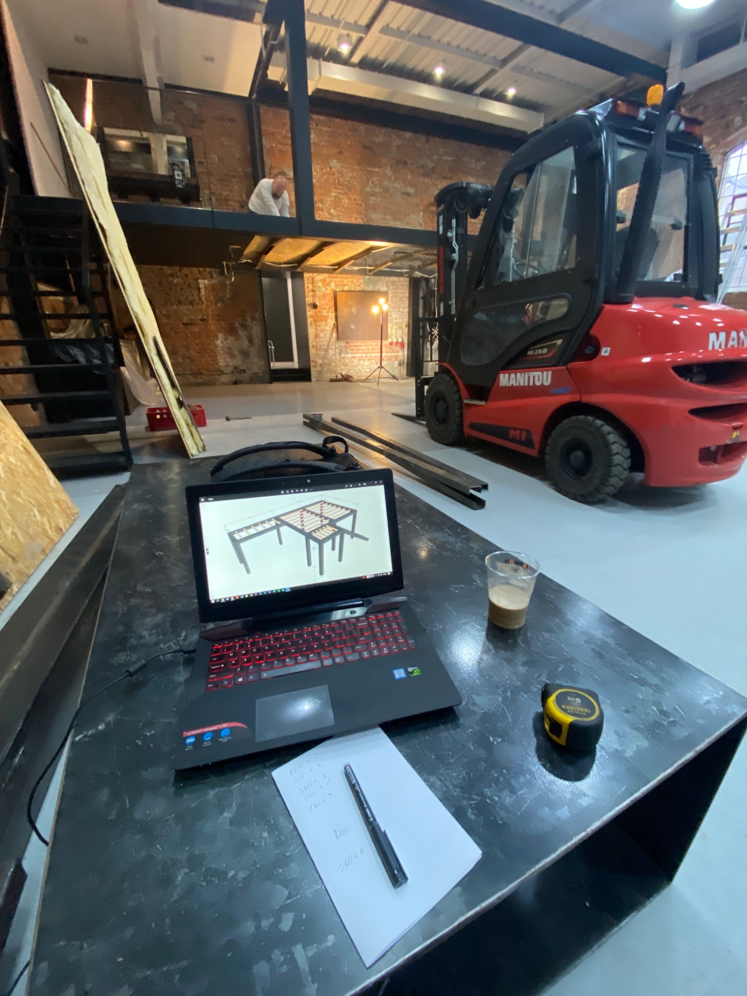Widok z góry na laptop z wizualizacją 3D konstrukcji metalowej, notatki, miarkę i napój na czarnym stole warsztatowym, w tle wózek widłowy i pracownik na antresoli w surowym wnętrzu hali.
