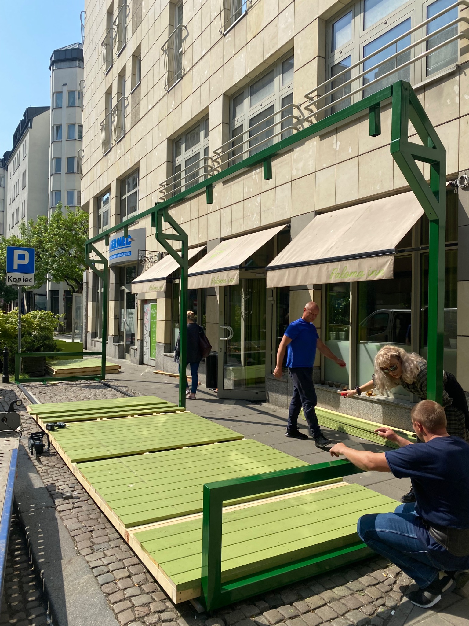 Montaż zielonej konstrukcji z metalu i drewna na chodniku przed budynkiem, prawdopodobnie elementu wyposażenia restauracji lub kawiarni.
