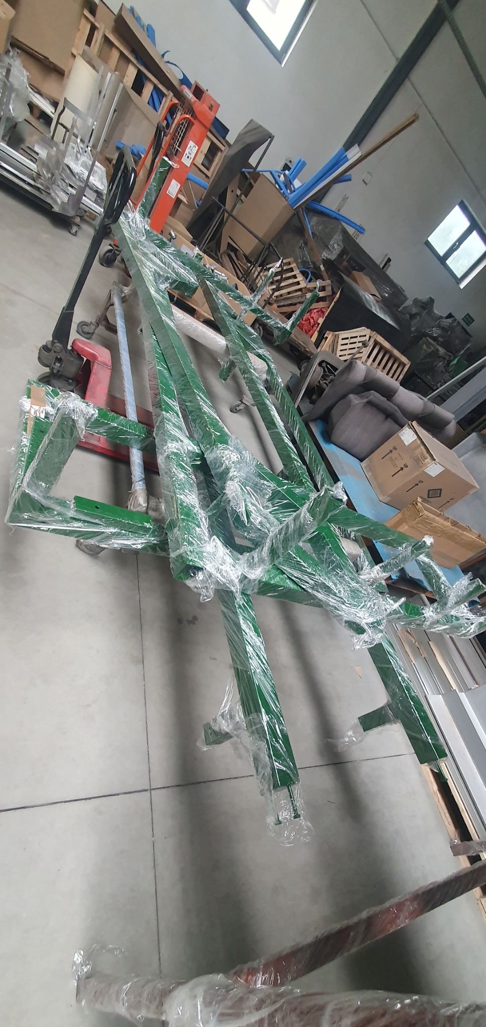 Elementy konstrukcji stalowej w kolorze zielonym, owinięte folią, przygotowane do transportu w Zielonce, widoczne w tle palety i wózek widłowy.