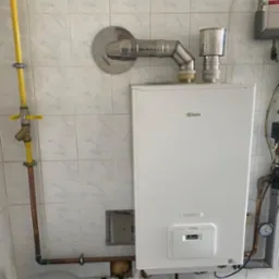 Biały wiszący piec gazowy Beretta Power X z podłączeniami gazowymi i wodnymi, zamontowany na ścianie wyłożonej marmurkowymi płytkami, z widocznym niebieskim pojemnikiem pod spodem.